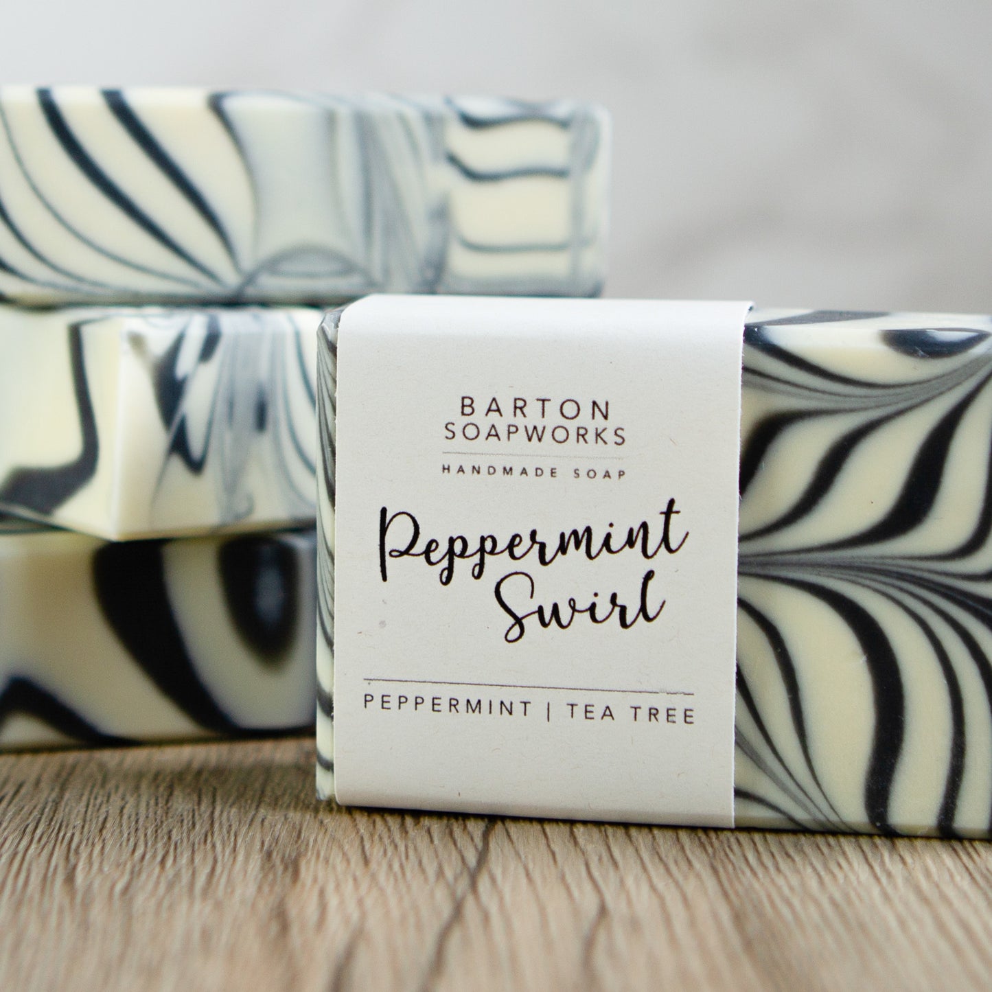 In the foreground is a bar of soap with a black and white swirled design. It has a cigar label on it which says Barton Soapworks, Handmade Soap, Peppermint Swirl, Pepperming and Tea Tree. In the background are 3 other bars stacked on top of one another. 